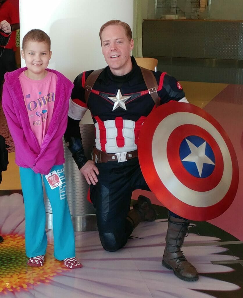 Sterling Bailey, founder of Cap for Kids, in a detailed Captain America costume kneeling next to a smiling young female cancer patient.