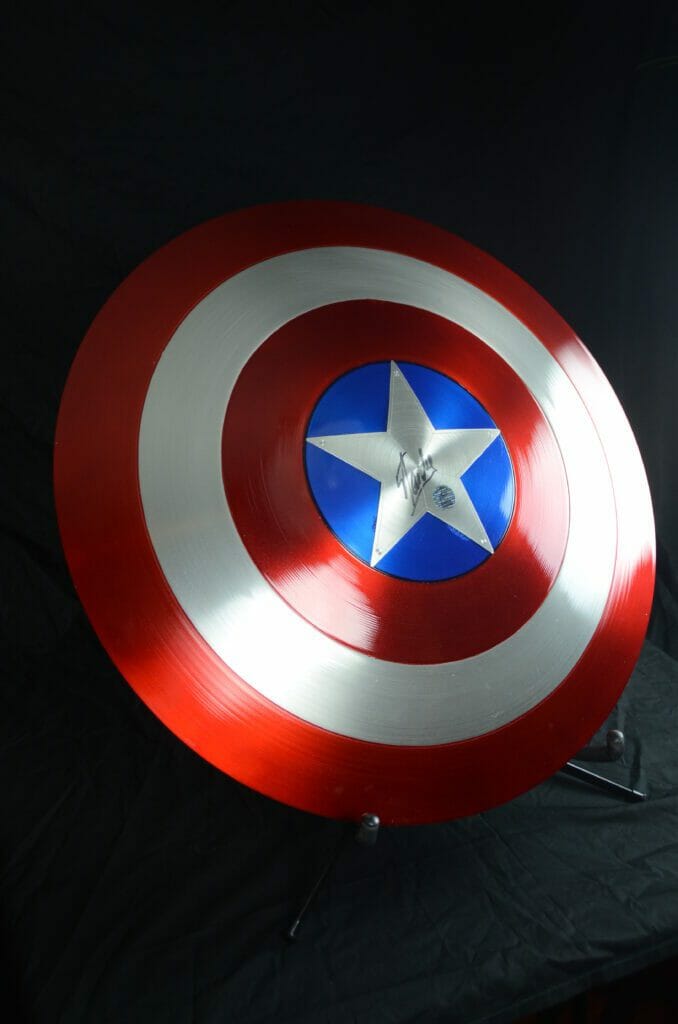 A Captain America shield photographed against a black background. The shield has Stan Lee's autograph in the center.